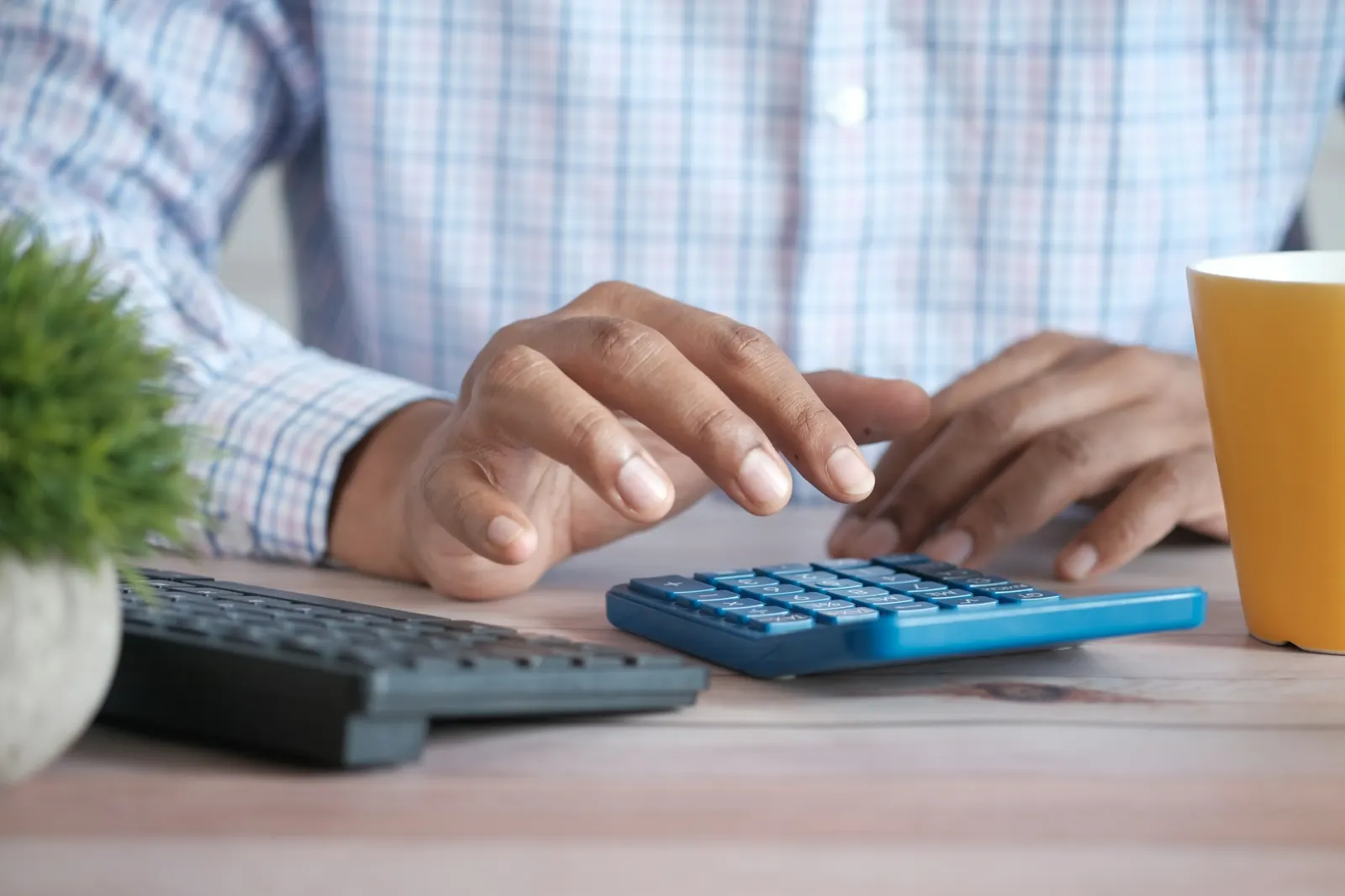 Hands working a calculator beside a keyboard and a yellow coffee mug on a bright desk