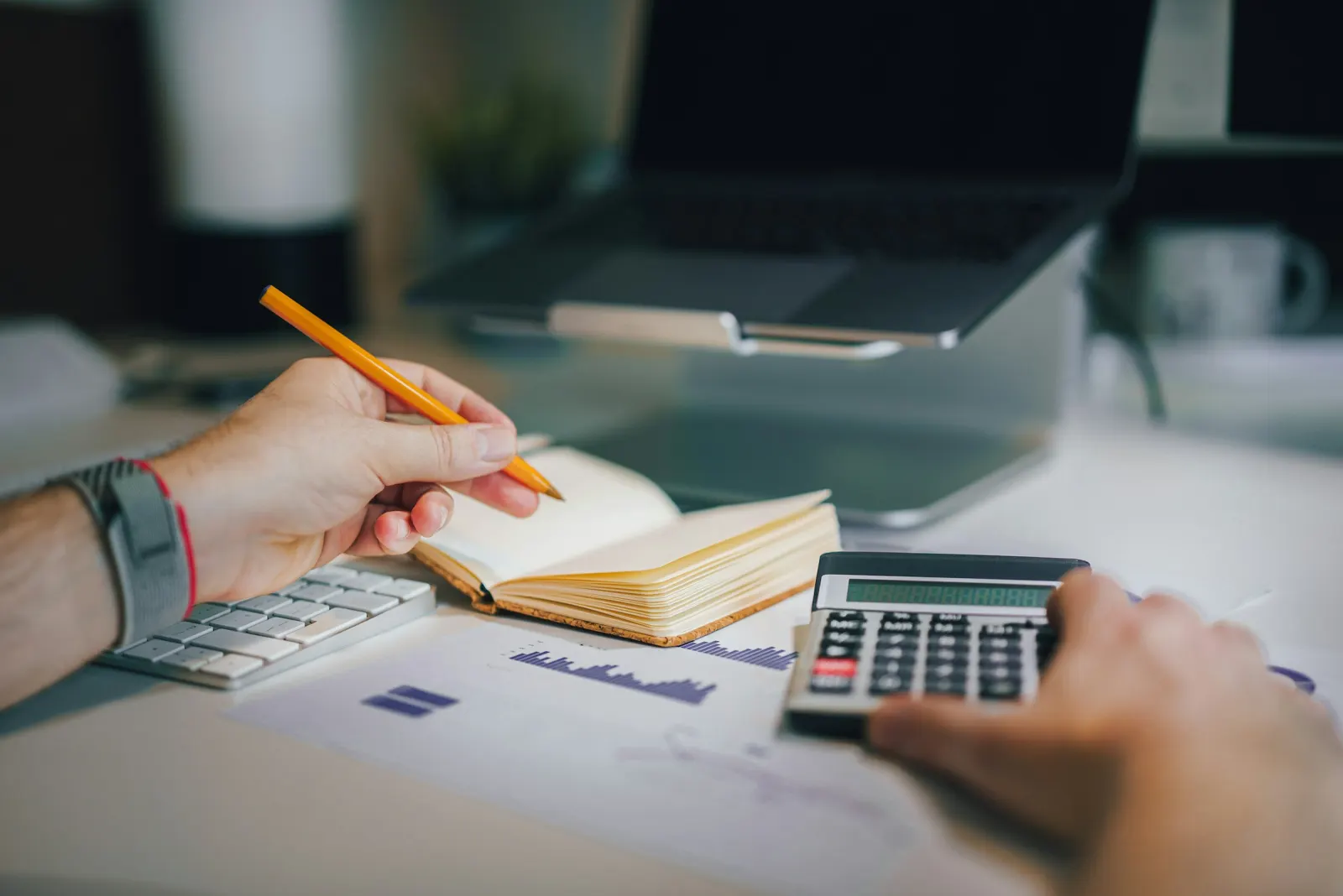 A person making notes in a notebook next to a calculator and printed figures on a white desk
