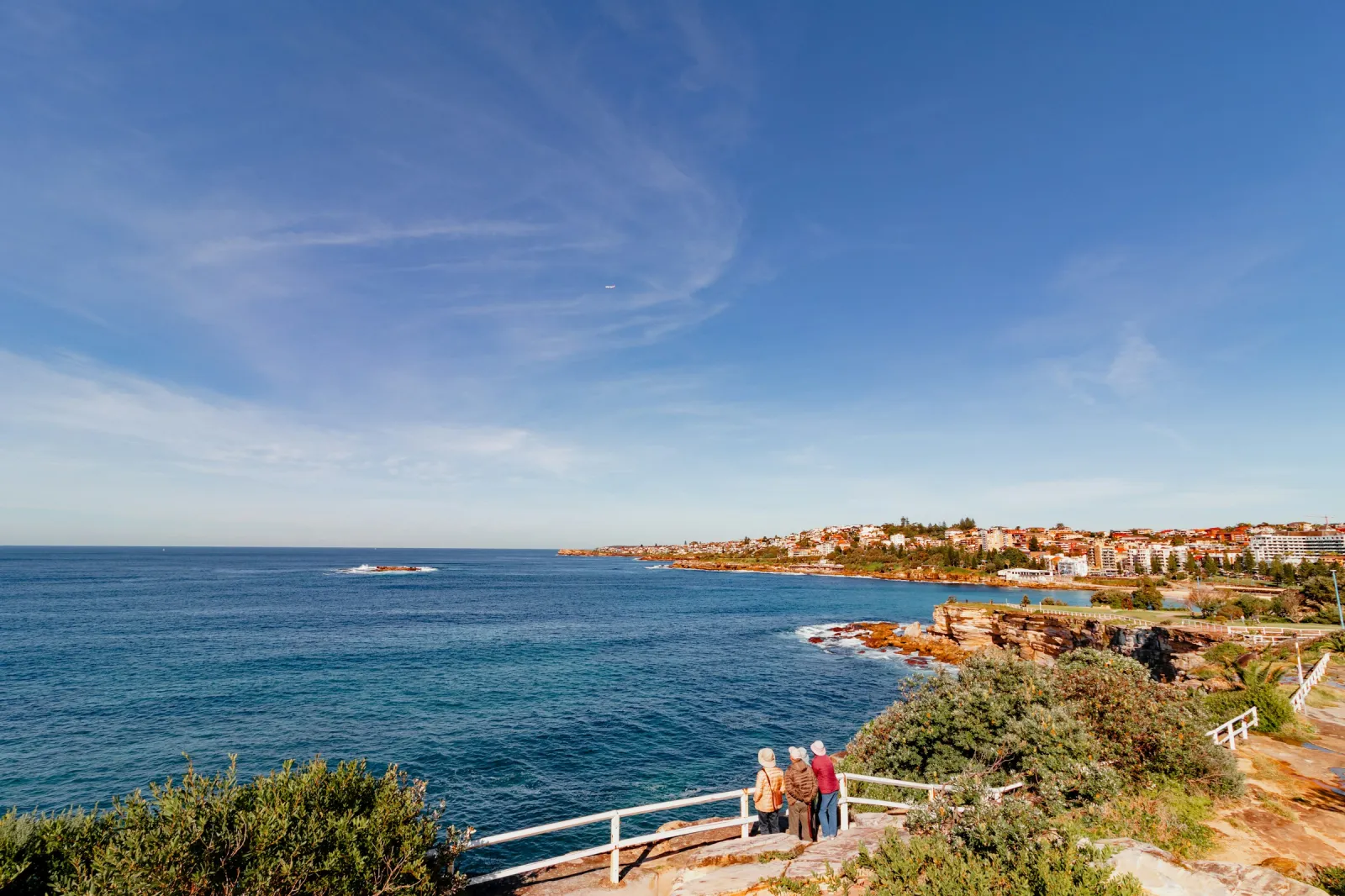 Two people at the railing of the coastal walk at Ben Buckler Point, North Bondi, looking out over a deep blue sea on a clear, sunny day