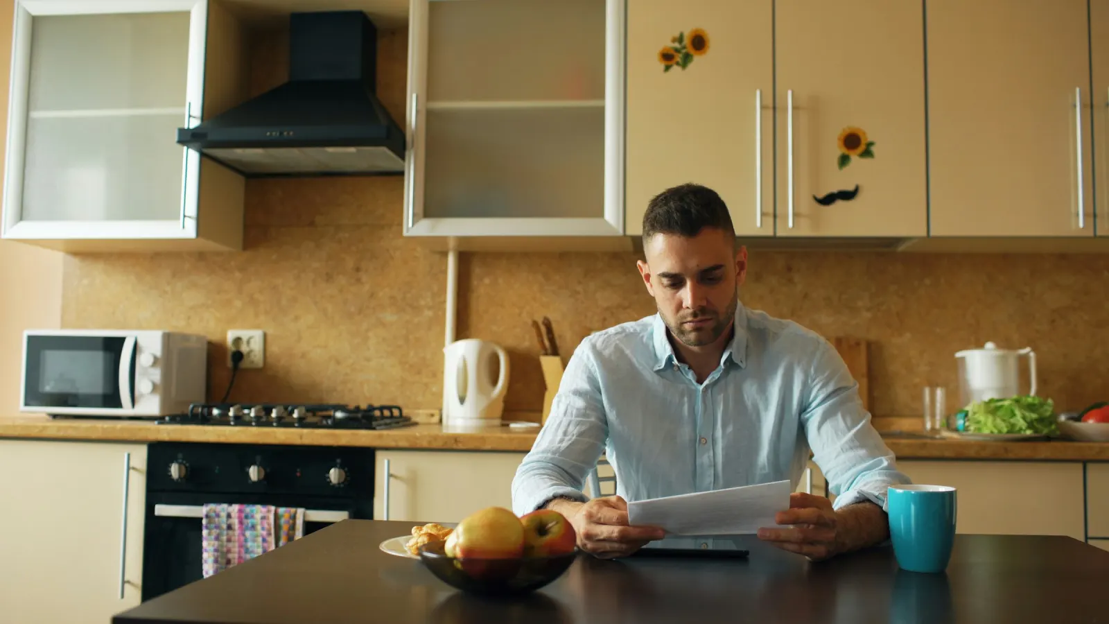 A man reading a letter at his kitchen table with a mug of coffee and a bowl of fruit beside him